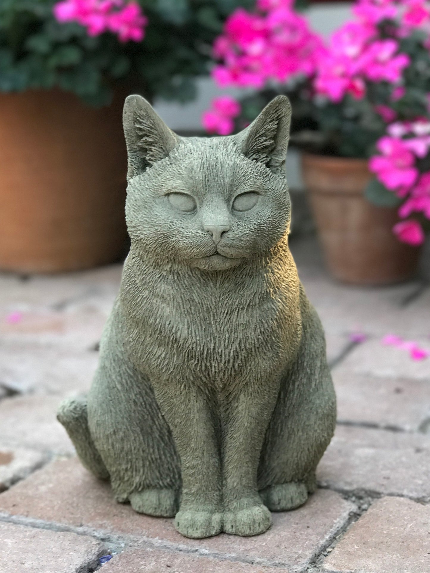 Katten Trisse – handgjuten betongkatt sittande i trädgård framför rosa blommor.