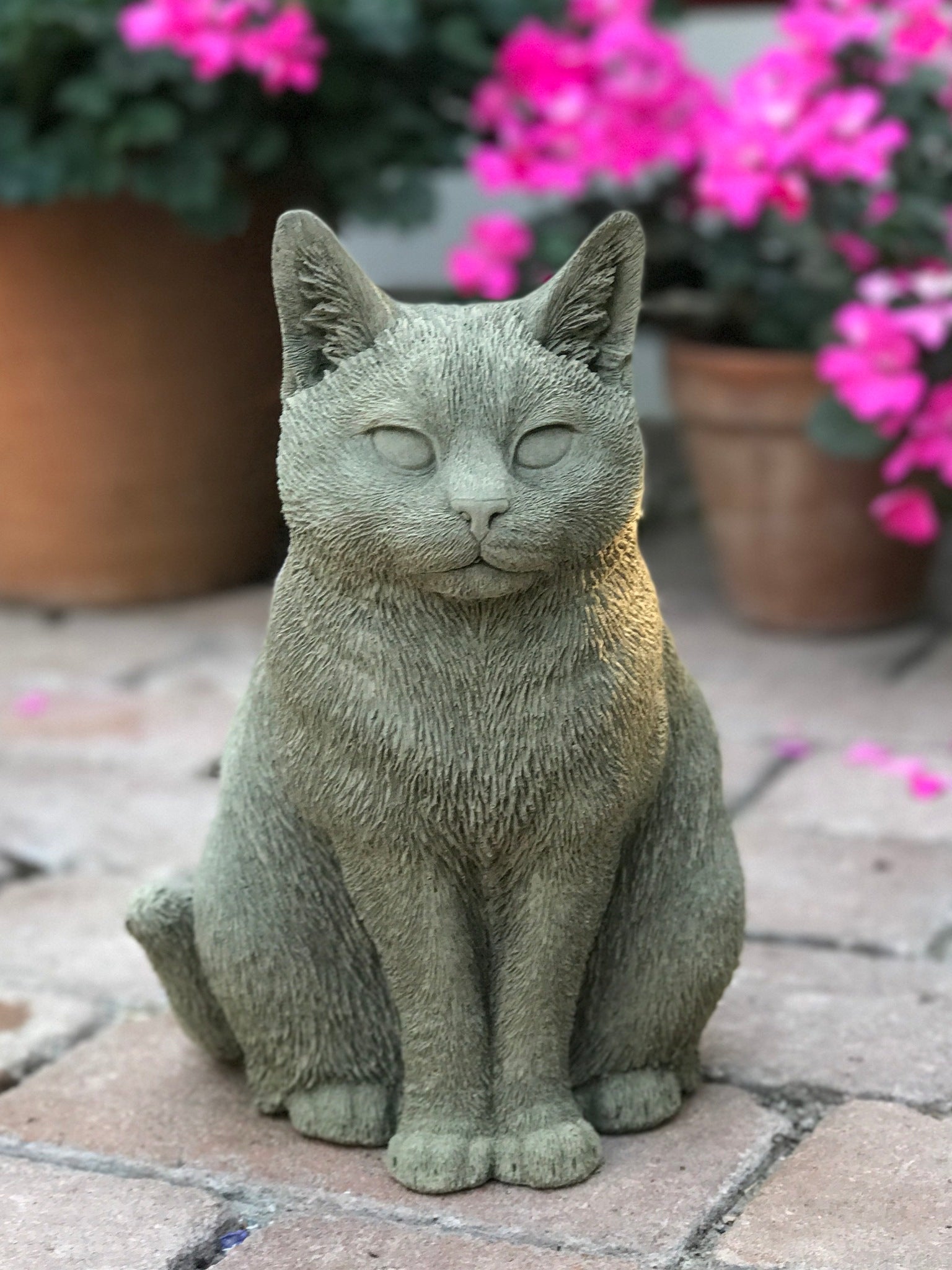 Katten Trisse – handgjuten betongkatt sittande i trädgård framför rosa blommor.
