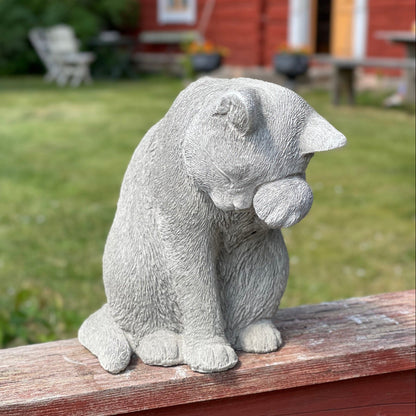 Betongkatt Selma som tvättar sig, placerad på en träregel utomhus